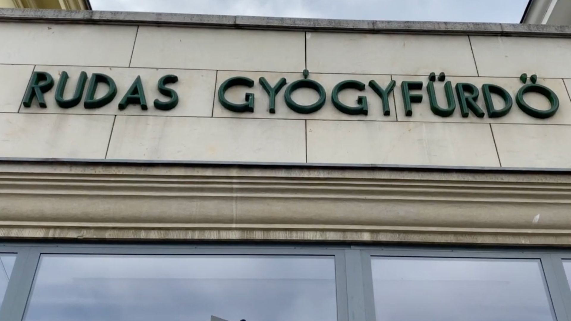 Close-up of the entrance sign for Rudas Gyógyfürdő, a historic thermal bath in Budapest, with green lettering on a beige stone facade.