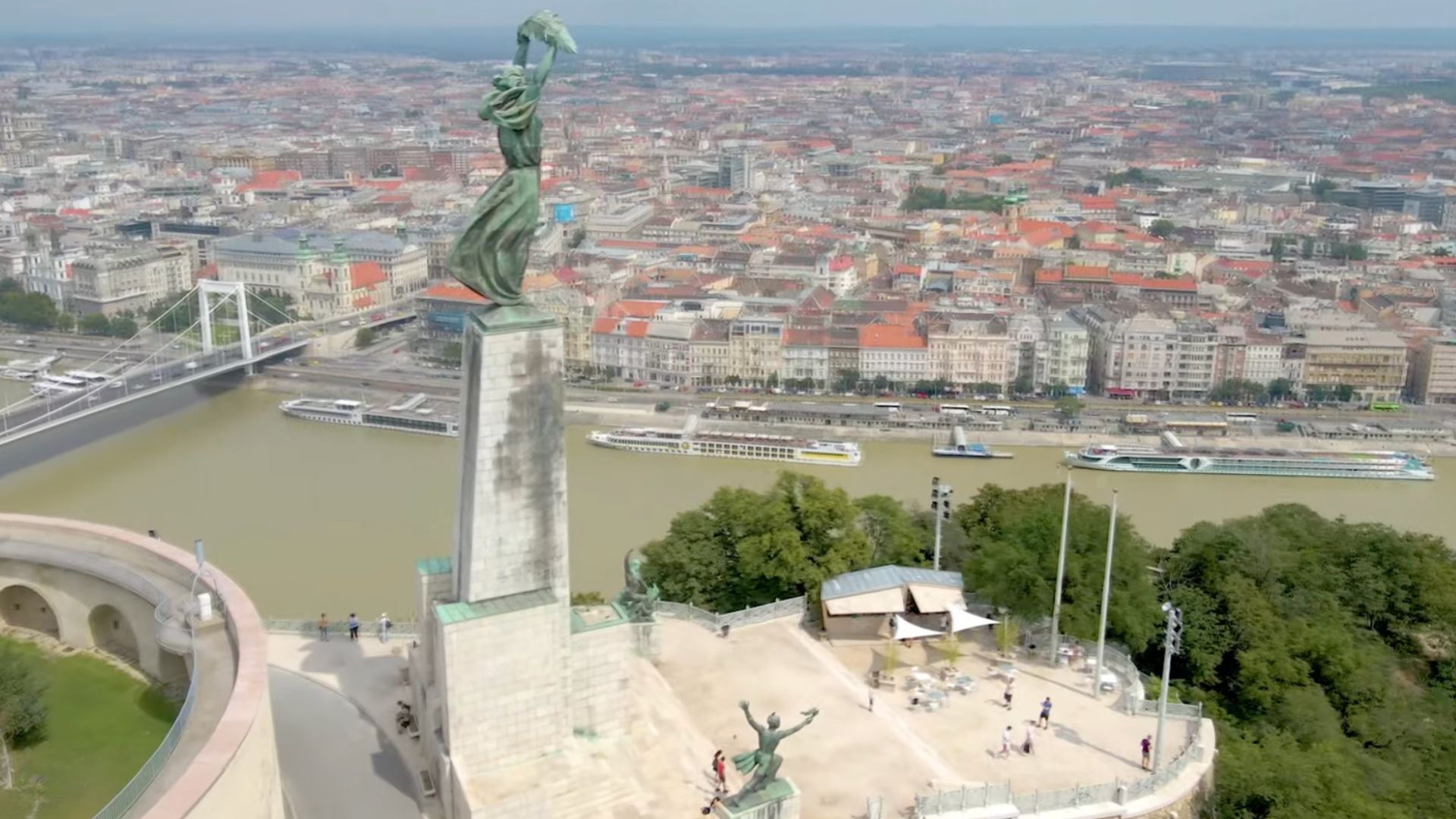 Aerial view of the Liberty Statue at the Citadella on Gellért Hill in Budapest, overlooking the Danube River and city skyline. The Elizabeth Bridge and riverboats are visible in the background.