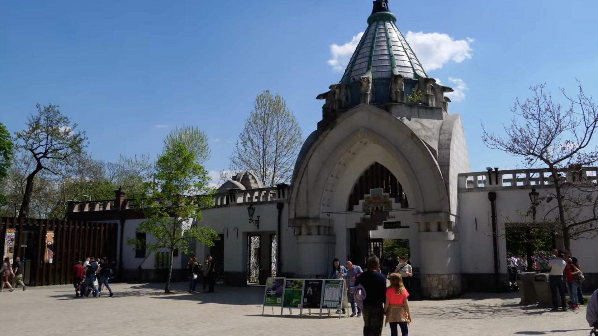 Entrance to the Budapest Zoo and Botanical Garden with a distinctive archway and spire, surrounded by trees on a sunny day with visitors walking by.