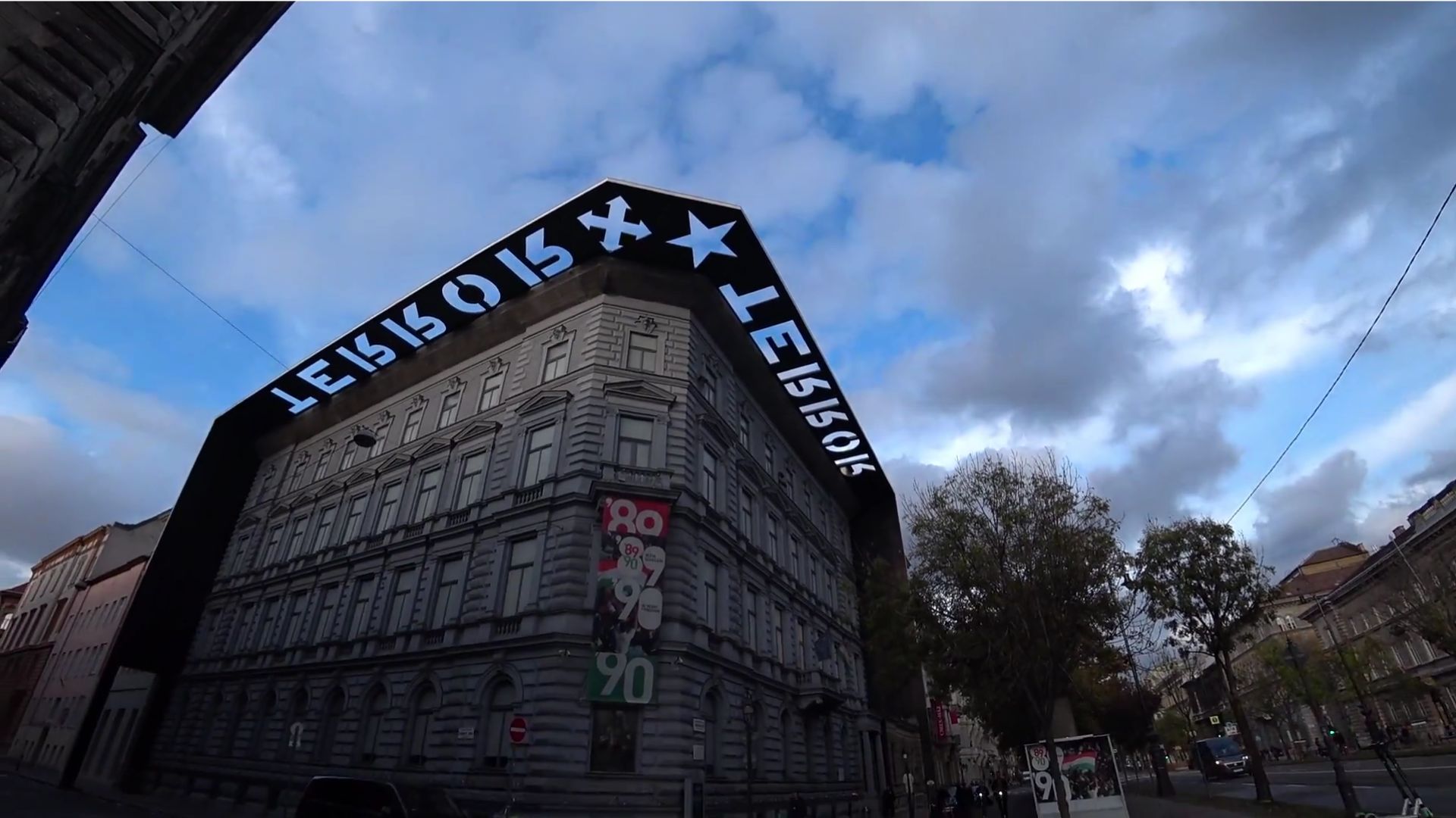 Exterior view of the House of Terror Museum in Budapest, showing the building's dark overhanging sign and architectural details under a cloudy sky.