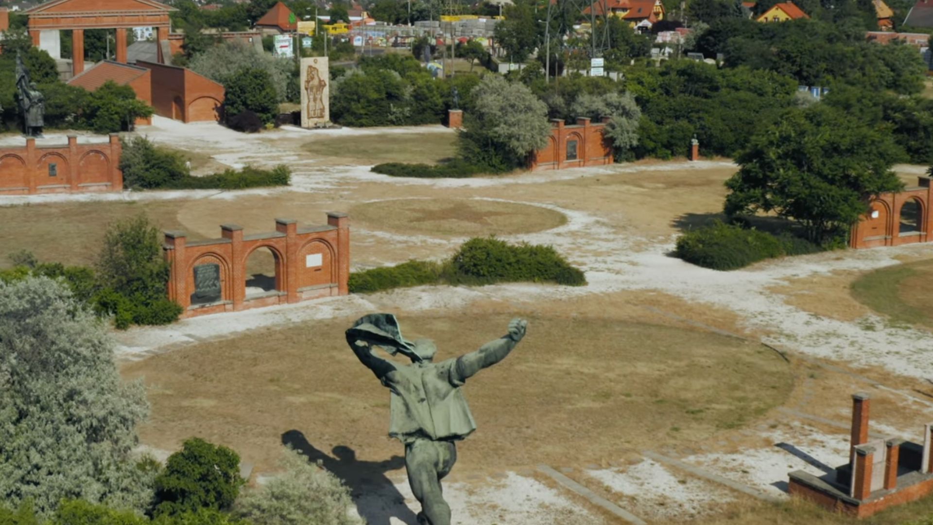 Aerial view of Memento Park in Budapest, showing scattered Soviet-era statues and red brick structures in an open, dry landscape.