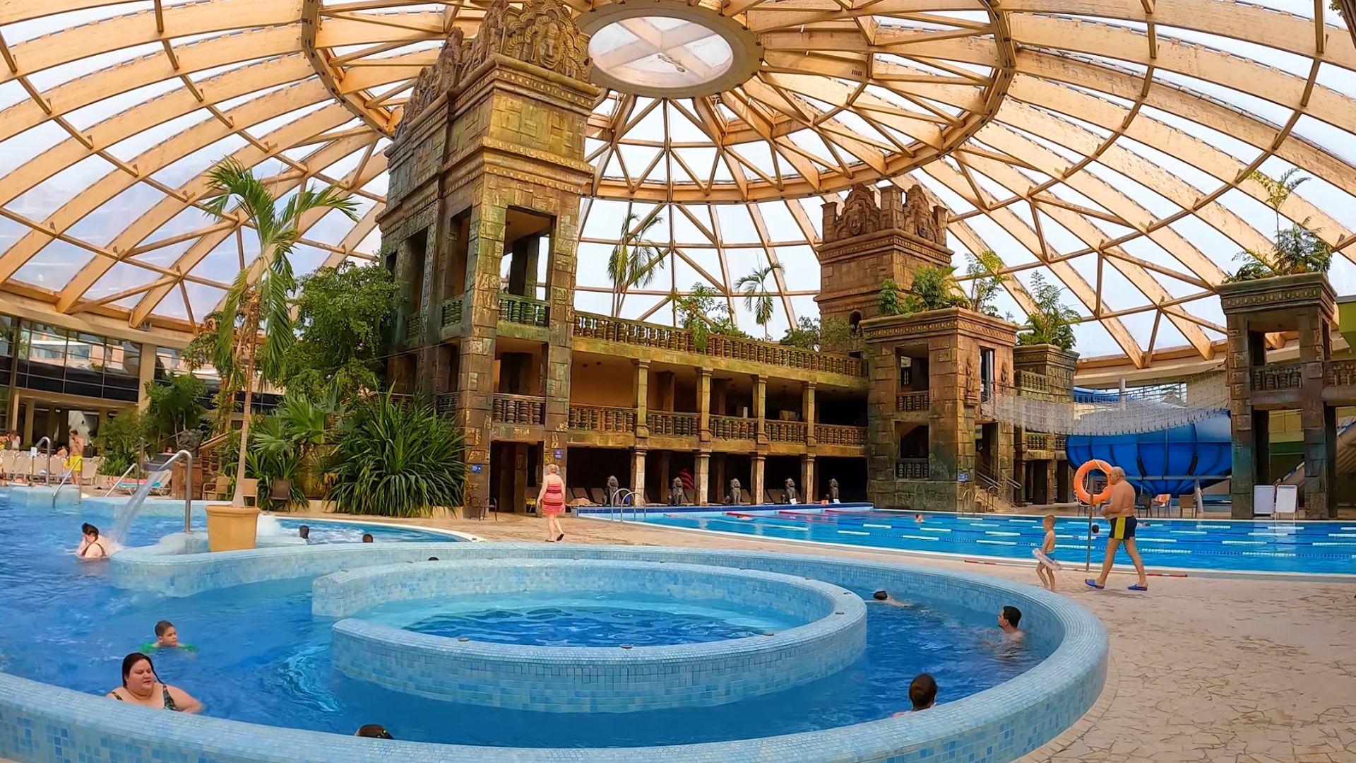 Aquaworld Aquapark in Budapest featuring a Mayan-themed indoor pool area with circular pools, towering structures, tropical plants, and a glass dome ceiling. People are swimming and enjoying the water.