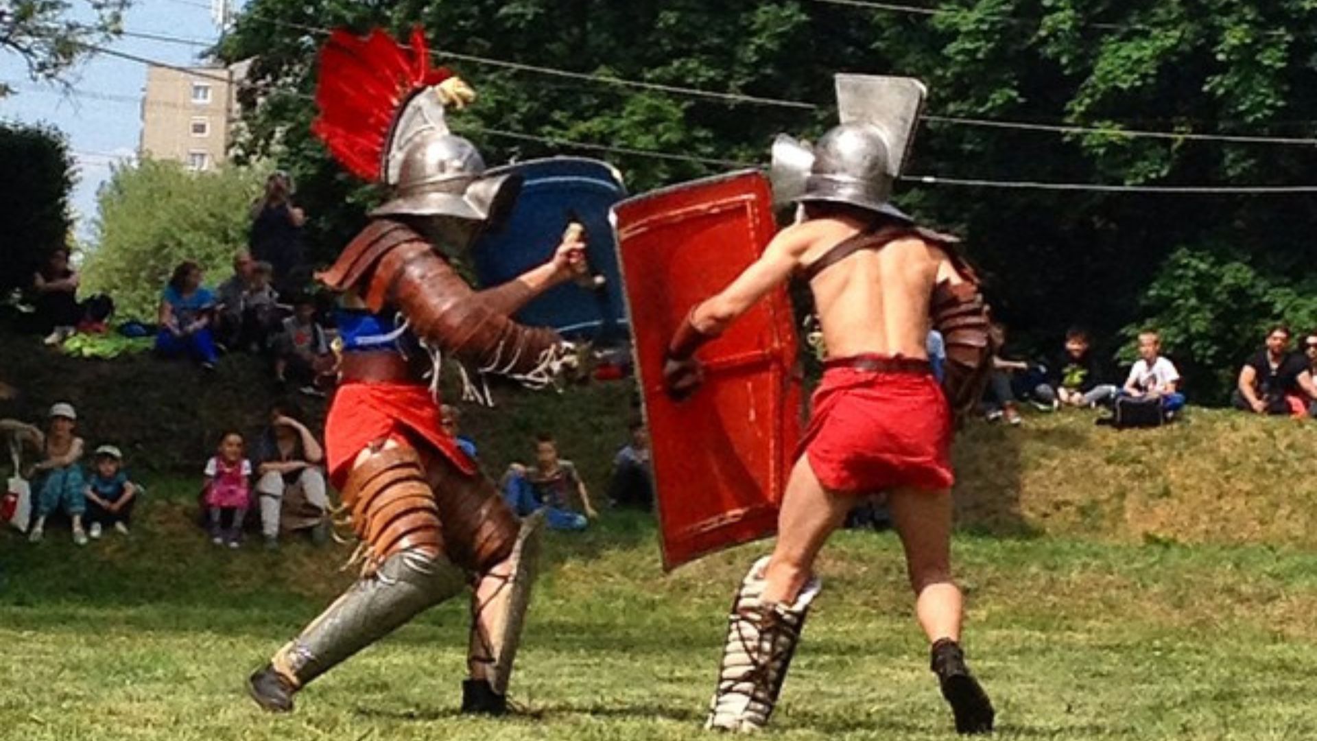 Gladiator reenactment at Aquincum with two performers in Roman armor and red attire battling in front of a seated audience outdoors.