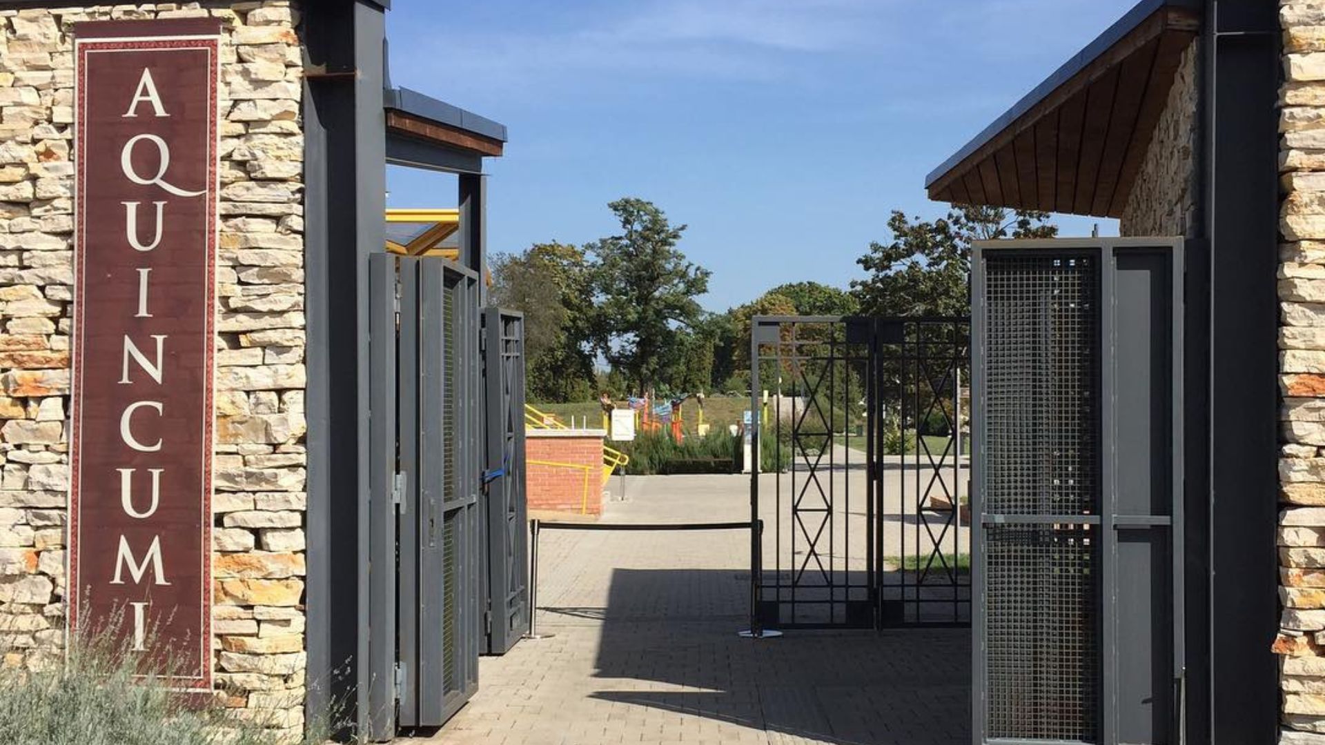 Entrance to the Aquincum Museum, featuring a modern gate with a stone wall and large maroon sign, surrounded by trees and a sunny sky.