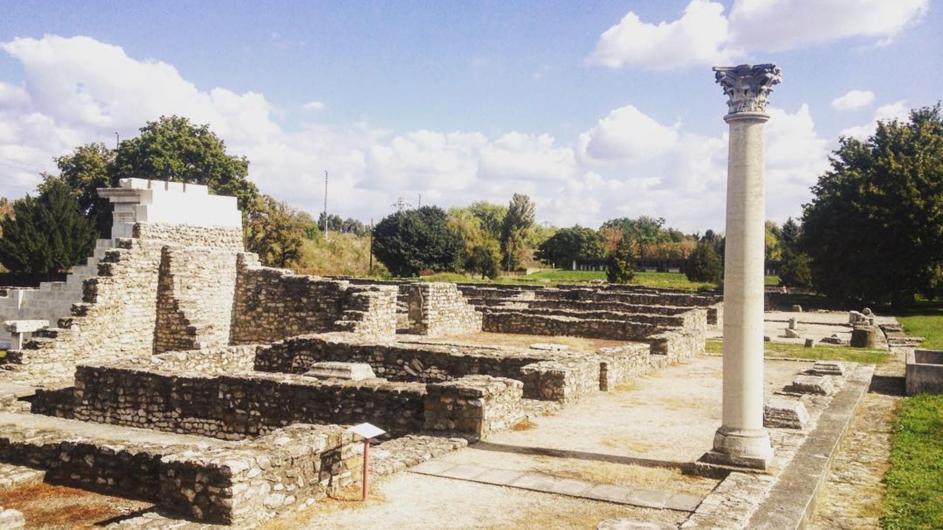 Expansive Roman ruins at Aquincum, with stone walls, a standing column, and a grassy landscape under a bright blue sky.